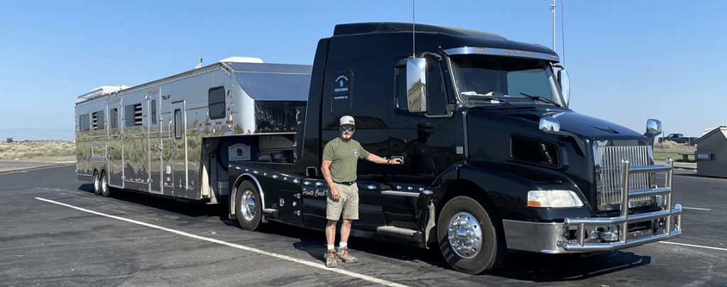 Richard standing beside a large black semi-truck hauling a shiny silver multi-horse trailer in a sunny, dry landscape.