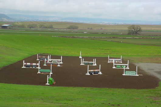 High-angle view of an outdoor jumping arena with multiple horse jumps set up on dark loam soil footing, surrounded by green hills.