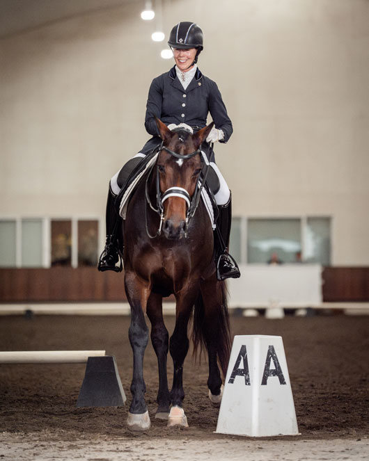 Equestrian rider Mary Burke competing on Brody, a bay Hanoverian gelding, performing a dressage test at the A marker in an indoor arena.