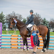 A female rider on a bay horse holding a championship ribbon alongside a trainer in front of a colorful butterfly jump.