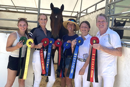 A group of five smiling riders and trainers at Burkeridge Farm holding several first, second, and third-place award ribbons in front of a horse