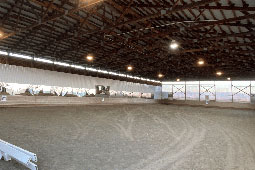 Inside view of a large indoor horse riding arena with a dirt floor, wooden roof trusses, and mirrors along the wall.